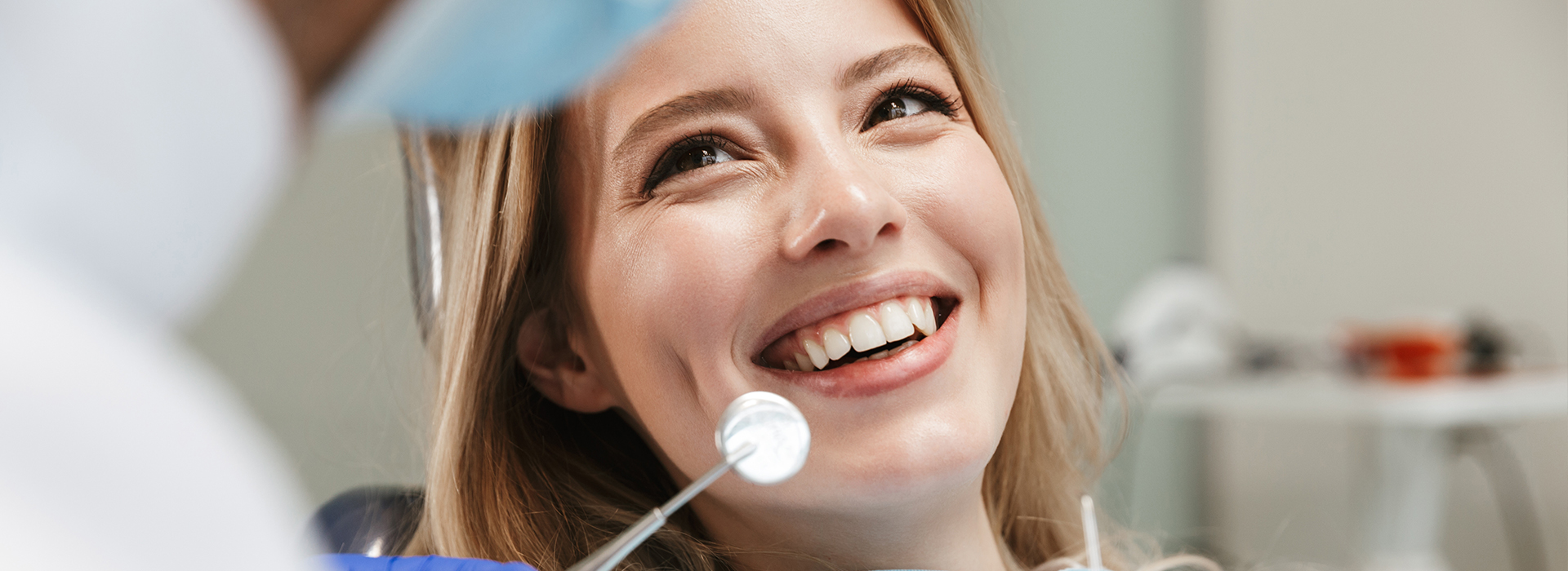 The image shows a young woman sitting in a dental chair with a smile on her face, looking at someone outside the frame, while a dentist works on her teeth.