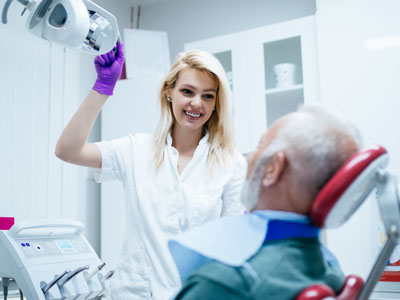A woman in a white coat stands next to an elderly man in a chair with medical equipment around him, holding a device above his head.